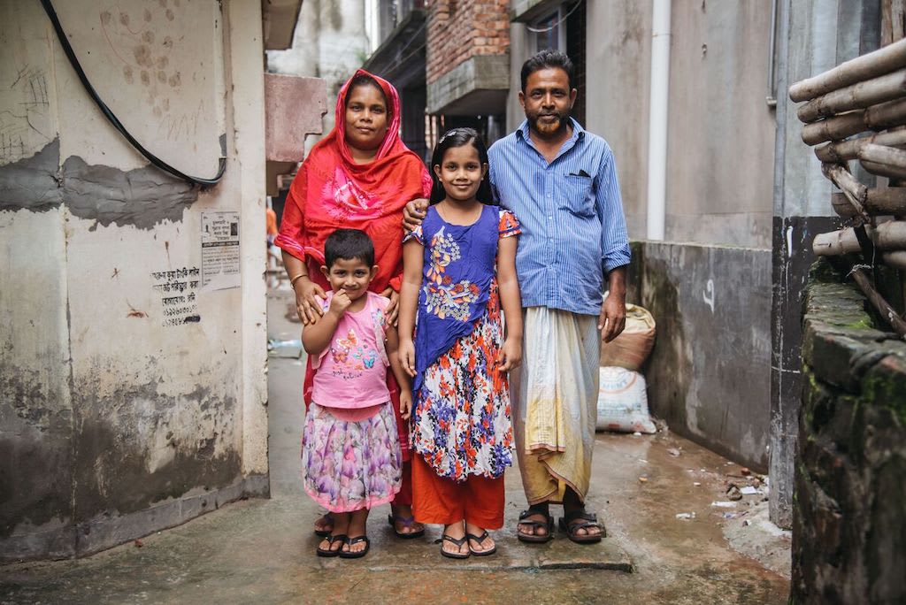 Mim is wearing an orange and purple dress. She is with her mother, wearing red, father, wearing a blue shirt and yellow pants, and her younger sister, wearing pink. They are standing outside their home with their arms around each other.